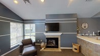 This single-floor community space is thoughtfully designed to provide a comfortable and inviting environment for residents and visitors alike. The layout encompasses distinct areas that blend functionality with style.

The main lounge area makes a strong first impression with soaring ceilings, a striking floor-to-ceiling stone fireplace featuring a rustic wood mantel, and an elegant chandelier overhead. Large French doors with dark trim frame views of the outdoors, filling the room with natural light. Comfortable seating, including plush sofas and wingback chairs, creates a relaxed and refined atmosphere.

A secondary lounge area offers a cozier setting, highlighted by a tile-surround fireplace topped with a classic white mantel and a wall-mounted television. Bold slate-blue walls accented with crisp white horizontal stripes give the room a contemporary character, while carpeted flooring and leather seating add warmth and comfort. Windows with blinds provide a connection to the surrounding landscape.

The reception area features a tiled wraparound desk with a white countertop, vaulted ceilings, and a practical kitchenette in the background. Neutral grey walls and tasteful wall art contribute to a clean, professional aesthetic.

Throughout the space, a cohesive color palette, textured finishes, and carefully chosen furnishings work together to create an atmosphere that is both welcoming and polished.