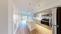 This thoughtfully designed 2-bedroom, 2-bathroom single-floor residence offers a functional and modern layout with clean finishes throughout. The open-concept kitchen serves as the heart of the home, featuring gray shaker-style cabinetry, a full suite of stainless steel appliances including a range, microwave, dishwasher, and refrigerator, and a central island with a white countertop ideal for dining or meal prep. Pendant lighting and a track light fixture add warmth and style overhead. Light hardwood flooring extends throughout the living areas, complementing the neutral white walls and creating a bright, airy ambiance. The bedrooms are comfortably sized, with the primary bedroom offering a dedicated closet equipped with wire shelving and hanging rods for organized storage. Natural light filters in through well-placed windows in the bedrooms. Both bathrooms are finished with hexagonal tile flooring, gray vanities topped with granite counters, and framed mirrors. One bathroom includes a full tub-shower combination with white subway tile surround. A hallway connects the rooms efficiently, and additional closet space with louvered doors provides practical storage. The overall aesthetic is clean, contemporary, and move-in ready.