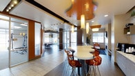 This single-floor amenity space showcases a contemporary design with a striking visual identity. The centerpiece of the room is an eye-catching, organically shaped drop ceiling panel finished in a bold burnt orange, complemented by elegant cylindrical pendant lights that cast a warm, inviting glow throughout the space. The ceiling design creates a dynamic focal point that draws the eye upward and adds architectural depth.

A sleek, curved white bar counter takes center stage, surrounded by modern amber-tinted acrylic bar stools with chrome bases, lending a stylish and cohesive color palette to the room. The flooring transitions between large-format light tiles and darker sections, subtly defining different zones within the open layout.

Rich dark wood columns and orange wall panels frame the space and reinforce the warm, energetic color scheme, while large windows allow ample natural light to filter in, brightening the interior and offering views of the surrounding greenery.

Toward the rear, a dedicated game area featuring a billiards table is visible, adding a recreational dimension to the space. Additional seating areas with lounge chairs provide comfortable spots for casual conversation. The overall ambiance is lively yet refined, making this a well-appointed space designed for both socializing and leisure.