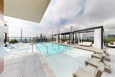 Located in the heart of Midtown Atlanta, this rooftop amenity level at 1081 Juniper St NE offers an elevated outdoor experience with a striking resort-style pool as its centerpiece. The generously sized swimming pool is finished in crisp aqua tile and is flanked by rows of modern white chaise lounges, ideal for sunbathing or relaxing against the backdrop of the Atlanta skyline. Dramatic pergola structures draped with flowing white curtains provide shaded cabana-style retreats with plush outdoor seating, creating a sense of private luxury amid the open sky. Multiple lounge areas are thoughtfully arranged across the expansive tile deck, including a covered seating zone with contemporary furniture, a fire feature, and raised planters filled with lush greenery that add a natural contrast to the building's warm terracotta and neutral paneling. An outdoor dining area with a round white table and modern chairs sits adjacent to floor-to-ceiling glass doors that connect to an interior amenity space. Panoramic views of the Midtown Atlanta skyline, including iconic high-rises, are visible from virtually every angle of the deck, making this rooftop an exceptional setting for both relaxation and social gatherings.