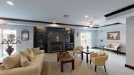 This Club Room is a well-appointed communal gathering space designed with both comfort and elegance in mind. The room's most striking architectural feature is a floor-to-ceiling dark granite fireplace wall, flanked by illuminated display niches that add depth and visual interest. The fireplace serves as a natural focal point, anchoring a generously sized seating arrangement of cream-toned sofas and armchairs paired with rich wooden coffee and side tables, all set atop plush neutral carpeting. Decorative dark wood ceiling beams frame a tray ceiling overhead, complemented by a ceiling fan with integrated lighting and recessed can lights that create a warm, layered ambiance. Bold dark wood crown molding and baseboards run throughout, providing a polished contrast to the soft blue-gray walls. Natural light flows in through glass-paneled exterior doors that offer views of an outdoor area beyond. A secondary seating nook features a pair of upholstered chairs, a petite side table, and framed wall art, providing a quieter retreat within the larger space. The entrance is defined by elegant dark wood-framed glass double doors, lending a refined, welcoming character to the room as a whole.