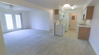 This single-floor space offers an open-concept layout that seamlessly connects the living area to the kitchen. The living area is finished with light-colored wall-to-wall carpeting, creating a neutral and versatile backdrop. Bright white walls throughout contribute to an airy, clean aesthetic.

The kitchen is positioned at the far end of the open space and transitions to tile flooring. It features white cabinetry, including upper wall-mounted cabinets with a wood-toned accent cabinet above, providing functional storage. The kitchen is equipped with a white electric range and a full-sized refrigerator, alongside an over-the-range microwave or ventilation unit. A pendant light fixture hangs overhead, adding a focal point to the kitchen area.

A doorway visible near the kitchen leads to an additional room or hallway, which appears to include an electrical panel, suggesting utility access nearby. A second door is visible along the back wall of the living area. The overall ambiance is clean, bright, and practical, offering a straightforward and functional living environment.