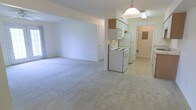This single-floor space offers an open-concept layout that seamlessly connects the living area to the kitchen. The living area is finished with light-colored wall-to-wall carpeting, creating a neutral and versatile backdrop. Bright white walls throughout contribute to an airy, clean aesthetic.

The kitchen is positioned at the far end of the open space and transitions to tile flooring. It features white cabinetry, including upper wall-mounted cabinets with a wood-toned accent cabinet above, providing functional storage. The kitchen is equipped with a white electric range and a full-sized refrigerator, alongside an over-the-range microwave or ventilation unit. A pendant light fixture hangs overhead, adding a focal point to the kitchen area.

A doorway visible near the kitchen leads to an additional room or hallway, which appears to include an electrical panel, suggesting utility access nearby. A second door is visible along the back wall of the living area. The overall ambiance is clean, bright, and practical, offering a straightforward and functional living environment.