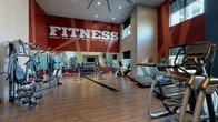 Located at 10501 Landing Way in Miamisburg, Ohio, this fitness center is a purpose-built exercise facility designed to energize and motivate. The space is defined by its impressively tall ceilings, which lend an open, expansive feel throughout the room. Bold graphic murals in deep red and vibrant yellow dominate the walls, emblazoned with motivational words such as "FITNESS," "workout," "push yourself," "endurance," and "stamina," creating a dynamic and high-energy atmosphere.

The flooring consists of durable hardwood-style planks that run across the entire space, providing a clean and functional surface for all types of workouts. Large floor-to-ceiling mirrors line key sections of the walls, visually expanding the space and allowing users to monitor their form during exercise.

The facility is comprehensively equipped with a wide range of cardio machines, including treadmills, ellipticals, stair climbers, and stationary bikes. A dedicated strength training area features a multi-station cable machine, a dumbbell rack with a full set of free weights, medicine balls, and an adjustable bench. A flat-screen television is mounted in a built-in niche on one wall, accompanied by storage cubbies below.

Natural light enters through high clerestory windows, complemented by recessed ceiling lighting. A glass-paneled entry door with a "WORKOUT" decal provides a welcoming entrance to this well-appointed fitness space.