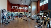 Located at 10501 Landing Way in Miamisburg, Ohio, this fitness center is a purpose-built exercise facility designed to energize and motivate. The space is defined by its impressively tall ceilings, which lend an open, expansive feel throughout the room. Bold graphic murals in deep red and vibrant yellow dominate the walls, emblazoned with motivational words such as "FITNESS," "workout," "push yourself," "endurance," and "stamina," creating a dynamic and high-energy atmosphere.

The flooring consists of durable hardwood-style planks that run across the entire space, providing a clean and functional surface for all types of workouts. Large floor-to-ceiling mirrors line key sections of the walls, visually expanding the space and allowing users to monitor their form during exercise.

The facility is comprehensively equipped with a wide range of cardio machines, including treadmills, ellipticals, stair climbers, and stationary bikes. A dedicated strength training area features a multi-station cable machine, a dumbbell rack with a full set of free weights, medicine balls, and an adjustable bench. A flat-screen television is mounted in a built-in niche on one wall, accompanied by storage cubbies below.

Natural light enters through high clerestory windows, complemented by recessed ceiling lighting. A glass-paneled entry door with a "WORKOUT" decal provides a welcoming entrance to this well-appointed fitness space.