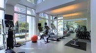 This contemporary fitness center offers a bright and open workout environment designed to support a full range of exercise activities. The space is defined by striking floor-to-ceiling windows that line the perimeter, flooding the interior with abundant natural light and providing views of lush palm trees and the surrounding urban landscape. The clean, white walls and polished light-toned flooring contribute to a crisp, modern aesthetic throughout.

The cardio zone is well-appointed with a row of treadmills and stationary exercise bikes, including high-end equipment suited for varied fitness levels. A stability ball and additional free weights and accessories are thoughtfully arranged along the windowed wall, making efficient use of the natural light. Large wall-to-wall mirrors on one side of the room visually expand the space and allow users to monitor their form during workouts.

The high ceilings enhance the airy, spacious feel of the facility, preventing any sense of confinement even during peak usage. The overall ambiance is clean, energizing, and professional, reflecting a commitment to providing a premium fitness experience. The layout is intelligently organized to allow for comfortable movement between equipment, making this fitness center a functional and inviting space for daily exercise routines.
