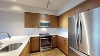 This well-designed single-floor residence offers a clean, contemporary aesthetic throughout. The open-concept living and kitchen area serves as the heart of the home, featuring warm-toned hardwood flooring that flows seamlessly from the living space to the kitchen. Large floor-to-ceiling sliding glass doors lead to a private balcony, flooding the living area with natural light and offering views of the surrounding cityscape and autumn foliage.

The kitchen is outfitted with flat-panel wood cabinetry in a rich walnut finish, white quartz countertops, and a full suite of stainless steel appliances, including a French door refrigerator, electric range, and over-the-range microwave. A white subway tile backsplash and track lighting complete the polished look. A peninsula provides additional counter space and casual seating.

The bedroom features plush carpeting, a generous window with exterior views, and access to a spacious walk-in closet equipped with multi-tiered wire shelving and hanging rods for ample storage. The bathroom is finished with wood-toned cabinetry, a white countertop vanity, a large mirror, and a sliding-glass enclosed walk-in shower. In-unit laundry is also conveniently included. Neutral tones and modern finishes create a cohesive, move-in-ready environment throughout.