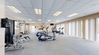 Located at 3901 Noyes Cir in Randallstown, Maryland, this fitness center offers a generously sized, single-floor exercise facility designed to accommodate a comprehensive workout experience. The space features a drop-panel ceiling with recessed fluorescent lighting that ensures bright, even illumination throughout, while soft neutral-toned walls contribute to a clean and welcoming atmosphere. Plush, patterned carpet tiles cover the floor, providing both comfort underfoot and a modern aesthetic.

The facility is equipped with a diverse selection of cardio machines, including elliptical trainers, upright and recumbent stationary bikes, and a treadmill. A multi-station cable weight machine and free weights, including wall-mounted foam dumbbells, cater to strength training needs. Fitness reference charts and a wall clock are posted for member convenience, and a wall-mounted flat-screen TV adds an entertainment element.

A standout feature of this space is the expansive wall of floor-to-ceiling windows and sliding glass doors fitted with vertical blinds, which flood the room with natural light and offer direct views of and access to an outdoor swimming pool and surrounding greenery. A small kitchenette area with cabinetry is also present. The overall ambiance is bright, functional, and inviting, making it a well-rounded community fitness resource.