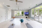 Located at 1040 Escalon Ave in Sunnyvale, California, this single-room yoga and fitness studio offers a clean, purpose-built environment designed to support wellness and mindful movement. The studio features light-toned hardwood-style flooring that spans the open, well-proportioned workout area, providing a bright and airy foundation for practice.

The space is painted in a calming slate-blue and white palette, contributing to a tranquil atmosphere ideal for yoga and exercise. Two ceiling fans help maintain comfortable airflow throughout sessions, while recessed lighting supplements the generous natural light that pours in through multiple large windows along the walls. A wall-mounted flat-screen television with a sound system provides options for guided workouts or ambient media.

Ample storage is available via a row of built-in closeted doors along one wall, offering organized space for equipment and accessories. A compact shelving unit in the corner provides additional storage for smaller items. The studio is equipped with yoga mats, blocks, and exercise balls, making it ready for immediate use.

A standout feature is the set of glass double doors with sidelights that open directly onto an outdoor patio area, seamlessly connecting the interior studio with a lush, tree-lined outdoor space complete with seating and umbrellas — ideal for extending practice outdoors.