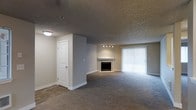 This single-floor space features an open living area finished with plush wall-to-wall carpeting that transitions to hard surface flooring near the entryway. The living room is anchored by a classic white-mantled fireplace, serving as a natural focal point of the space. A set of vertical blinds covers a large sliding glass door, which allows natural light to fill the room while providing access to the outdoors. Track lighting above the fireplace offers targeted illumination, complementing the flush-mount ceiling light near the entry. The textured popcorn ceiling adds a traditional character throughout. A white panel door leads to an additional room, suggesting a functional layout with defined, separate spaces. Neutral gray walls and understated finishes throughout give the space a clean, move-in-ready appearance. The open arrangement of the main living area allows for flexible furniture placement, making it adaptable to a variety of needs. Overall, the space presents a straightforward and practical design suited for comfortable everyday living.