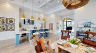 Located at 3625 Frankford Ave in Panama City, Florida, this beautifully designed single-floor space blends coastal charm with upscale modern aesthetics. The open-concept layout is anchored by soaring coffered ceilings that lend an airy, expansive feel throughout. Light hardwood flooring runs seamlessly across the entire space, complementing the bright white walls and trim.

A standout feature is the vivid turquoise bar-height island topped with a crisp white quartz countertop, surrounded by stylish barstool seating. Above it, a trio of bold gold pendant lights adds warmth and a touch of glamour. A beverage station and additional counter space line the adjacent wall, making this area ideal for gatherings and entertaining.

The lounge area is thoughtfully curated with rich leather armchairs, colorful accent pillows, and layered area rugs, creating an inviting and comfortable atmosphere. Coastal-themed artwork, including egret paintings and seascape prints, adorns the walls throughout, reinforcing the relaxed Florida aesthetic.

Large black-framed glass doors and an oversized garage-style window flood the interior with natural light and offer views of the outdoors. A dedicated entry foyer features dramatic vaulted paneled walls and additional coastal artwork, setting an elegant tone upon arrival. The overall ambiance is warm, refined, and distinctly coastal.