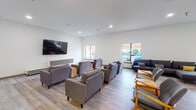 This single-floor communal facility offers a well-appointed, contemporary environment designed for social gathering and recreation. The main lounge area is generously sized, featuring light wood-toned flooring, crisp white walls, and recessed lighting throughout. Multiple seating arrangements are thoughtfully configured across the space, with plush grey upholstered armchairs and sofas accented by yellow throw pillows, complemented by distinctive log-slice side tables that add a natural, rustic touch. A large wall-mounted flat-screen television serves as a central focal point in the primary lounge zone, making it well-suited for group viewing or relaxed socializing. Several windows line the walls, allowing natural light to fill the interior and providing views of the surrounding exterior. An adjacent games room offers dedicated space for recreation, housing both a shuffleboard table and a ping-pong table on the same light hardwood flooring, providing a casual and engaging environment for interactive activities. A separate open area transitions into an additional lounge zone, distinguished by patterned carpet flooring, bold yellow ceiling accents, and exposed industrial-style ductwork, lending an eclectic, creative character to that section of the space. Together, the areas form a cohesive, versatile community hub designed to accommodate a variety of social and recreational needs.