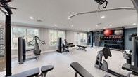Located at 2124 Gables Dr NE in Atlanta, GA, this single-floor fitness center offers a thoughtfully designed space for a full-spectrum workout experience. The layout is open and spacious, finished with neutral gray carpeting and smooth white walls accented by a bold navy blue feature wall that anchors the functional training zone. Recessed lighting throughout ensures bright, even illumination across the entire floor.

The strength training area is well-appointed with multiple cable machines, weight benches, a lat pulldown station, and selectorized equipment suited for a variety of muscle groups. A large mirror along one wall enhances the sense of depth and allows users to monitor their form. The functional training zone stands out with a fully stocked rack of kettlebells, medicine balls, and resistance tools, complemented by a heavy punching bag and suspension training anchors mounted to the wall.

Natural light flows in generously through multiple large, grid-paned windows and a glass-paneled exterior door, offering views of the surrounding greenery. The door provides direct outdoor access, adding convenience and ventilation. A water fountain is conveniently positioned near the exit corridor. The overall ambiance is clean, modern, and motivating — a professional-grade fitness environment designed to support diverse training goals.