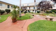 This multi-unit property is centered around a thoughtfully landscaped communal courtyard that serves as its defining feature. The stucco-clad buildings, finished in a neutral cream tone, frame the outdoor space on multiple sides, creating an enclosed and cohesive environment. Exterior staircases with metal railings provide access between levels, while large windows fitted with vertical blinds allow natural light to reach each unit.

The courtyard itself is generously sized and carefully maintained, featuring well-kept green lawns interspersed with curved, terracotta-colored concrete pathways that wind throughout the space. A classic tiered stone fountain serves as a central focal point, adding a touch of elegance to the outdoor area. Tall, mature palm trees and ornamental trees, including a striking purple-leafed variety, lend vertical interest and shade. Rounded, manicured shrubs, ornamental grasses, and mixed garden beds line the walkways, contributing to a layered, lush aesthetic. Natural boulders are placed as decorative accents within the lawn areas. The overall ambiance is tranquil and resort-like, with the landscaping creating a welcoming, park-style atmosphere throughout the shared outdoor space.