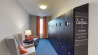 This single-floor space serves as a dedicated package and parcel locker room, thoughtfully designed to balance secure storage functionality with a welcoming ambiance. The room is dominated by a bank of tall, navy blue lockers equipped with individual digital keypad entry systems, providing secure and convenient access to stored items. The lockers are arranged in a multi-unit, floor-to-ceiling configuration that maximizes storage capacity along one wall.

Complementing the utilitarian purpose of the lockers, the space incorporates a comfortable seating area furnished with neutral upholstered armchairs accented with coral and orange throw pillows. A small side table with a warm-toned ceramic lamp adds a residential touch, creating a softened atmosphere within an otherwise functional space.

A window dressed with bold, rust-orange curtain panels introduces natural light and a pop of color, enhancing the room's visual warmth. The blue-toned carpeting ties cohesively to the color palette of the lockers, while a flush-mount ceiling light provides even overhead illumination. The overall design merges practical secure storage with an inviting, well-appointed aesthetic.
