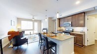 This single-floor residence showcases a thoughtfully designed open-concept layout that connects the kitchen and living areas into one cohesive, functional space. The kitchen serves as a central focal point, featuring rich dark-toned cabinetry that extends to the upper walls, complemented by granite countertops with warm, natural veining. A peninsula island with seating for multiple bar-height chairs provides both additional workspace and a casual dining option, illuminated by sleek pendant lighting overhead. Stainless steel appliances, including a gas range and built-in microwave, reinforce the modern aesthetic throughout the kitchen.

Dark hardwood-style flooring runs continuously through the space, grounding the design and providing a striking contrast against the light-toned walls and ceiling. A ceiling fan anchors the living area, which benefits from generous natural light streaming through large windows and a glass-paneled door. The living space accommodates a comfortable seating arrangement atop a patterned area rug, creating a warm, welcoming atmosphere. The overall design balances contemporary finishes with practical living features, resulting in a cohesive and stylish interior.