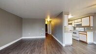 This single-floor residential unit offers a well-organized layout designed for comfortable everyday living. The open living area is generously sized, finished with durable hardwood-style flooring in a cool gray tone and accented by clean white baseboards. A half-wall partition separates the living space from the adjacent galley-style kitchen, which features white cabinetry with warm wood-trimmed edges, granite-pattern countertops, a full suite of white appliances including an electric range and refrigerator, and a built-in sink with ample counter space on both sides. Natural light filters into multiple rooms through windows fitted with vertical blinds, offering views of surrounding greenery. The bedrooms are carpeted for comfort, with neutral-toned walls and closet access. A full bathroom off the hallway includes a toilet, vanity with countertop, and hardwood-style flooring. A dedicated laundry area tucked within the hallway houses a stacked washer and dryer unit for added convenience. The hallway connects all areas of the unit efficiently, and the overall color palette of soft grays and warm neutrals lends a clean, move-in-ready aesthetic throughout.