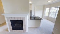 This single-floor residence offers a thoughtfully designed open-concept layout that seamlessly connects the living, dining, and kitchen areas. The living space is anchored by a classic tile-surround fireplace with a sleek black firebox insert, adding warmth and a focal point to the room. Soft, plush carpeting flows throughout the main living areas, contributing to a comfortable and inviting atmosphere. Crown molding along the ceiling adds a refined architectural touch, while neutral, light-toned walls create a bright and airy feel throughout.

The kitchen is defined by a peninsula-style breakfast bar featuring a striking dark granite countertop that contrasts elegantly with the white cabinetry and light surroundings. A stainless steel refrigerator anchors the kitchen, complementing the modern aesthetic. The peninsula also features an integrated sink, making it a highly functional workspace. A white paneled door provides access to additional storage or utility space within the kitchen area.

Natural light filters in through multiple windows, enhancing the open and spacious feel of the home. The seamless flow between the living and kitchen areas makes this space well-suited for both everyday living and entertaining.