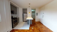 Located at 10 Marshall St in Irvington, NJ, this single-floor unit offers a well-designed layout with a blend of modern updates and classic architectural details. The kitchen is a standout feature, equipped with stainless steel appliances including a gas range, microwave, and refrigerator, complemented by contemporary gray cabinetry and clean white tile flooring. A central kitchen island with pendant lighting serves as both a functional workspace and a natural transition point into the living area. Throughout the main living spaces, warm parquet hardwood flooring adds a timeless character and visual richness to the interior. The living area is bright and open, featuring white walls and a window fitted with blinds that allows natural light to filter in. A dedicated entry area includes a utility panel and access to a walk-in closet, providing practical storage solutions. The bathroom is finished with marble-style wall and floor tiling, and includes a pedestal sink, mirrored medicine cabinet, and a glass-enclosed shower-tub combination, creating a clean and polished aesthetic. Baseboard heating is present throughout the unit, ensuring comfort year-round. Overall, the space presents a functional and updated living environment in a residential Irvington location.