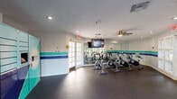This fitness center at Reserve at Lake Irene is a well-appointed, single-floor facility designed to support a full range of workout routines. The space is divided into distinct functional zones, each thoughtfully equipped and finished with a consistent teal, aqua, and navy color-banded design along the walls, lending a cohesive and energetic aesthetic throughout.

The spin studio is the centerpiece of the facility, featuring a row of professional-grade cycling bikes arranged on durable rubber flooring. A large wall-mounted flat-screen TV provides on-demand fitness content, while a full-length mirror along one wall amplifies the sense of space and allows riders to monitor their form. A ceiling fan overhead ensures comfortable airflow during intense sessions. French doors and surrounding windows invite ample natural light and offer views of the lush outdoor greenery.

An adjacent functional training area provides open floor space ideal for stretching, mat work, and balance training. A BOSU ball and medicine ball rack are available for varied workouts, and open-grid shelving units offer convenient storage for personal items and accessories.

The facility also includes a dedicated locker area outfitted with a bank of teal-colored smart lockers branded with the Reserve at Lake Irene logo, providing secure storage for residents. Recessed lighting throughout maintains a bright, clean atmosphere across all areas of the fitness center.