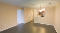 This single-floor residence offers a clean, open-concept layout that seamlessly connects the living, dining, and kitchen areas. Dark hardwood flooring runs throughout the main living space, providing a rich contrast against the warm, neutral-toned walls painted in a soft peach-beige palette. The kitchen features dark cabinetry paired with stainless steel appliances, including a built-in microwave and range, and is anchored by a raised peninsula with a sink — an ideal space for casual dining or meal prep. A pendant light fixture hangs above the peninsula, adding a refined focal point to the kitchen area. The open corridor leading toward the back of the residence reveals multiple doorways, suggesting additional rooms such as bedrooms or bathrooms, all accessible from the central hallway. White panel doors and white baseboards throughout lend a crisp, finished look to the interior. The layout is practical and well-proportioned, with a thoughtful flow between spaces. The overall ambiance is bright, clean, and move-in ready, offering a functional and welcoming living environment.
