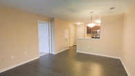 This single-floor residence offers a clean, open-concept layout that seamlessly connects the living, dining, and kitchen areas. Dark hardwood flooring runs throughout the main living space, providing a rich contrast against the warm, neutral-toned walls painted in a soft peach-beige palette. The kitchen features dark cabinetry paired with stainless steel appliances, including a built-in microwave and range, and is anchored by a raised peninsula with a sink — an ideal space for casual dining or meal prep. A pendant light fixture hangs above the peninsula, adding a refined focal point to the kitchen area. The open corridor leading toward the back of the residence reveals multiple doorways, suggesting additional rooms such as bedrooms or bathrooms, all accessible from the central hallway. White panel doors and white baseboards throughout lend a crisp, finished look to the interior. The layout is practical and well-proportioned, with a thoughtful flow between spaces. The overall ambiance is bright, clean, and move-in ready, offering a functional and welcoming living environment.