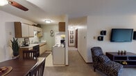 This single-floor unit offers a practical and comfortable layout across several well-defined living spaces. The kitchen is configured in a galley style, featuring cream-toned cabinetry with wood trim accents, a double stainless steel sink, electric coil range, refrigerator, dishwasher, and tile flooring. A central support column partially separates the kitchen from the adjoining areas, lending subtle architectural definition to the space.

The dining area accommodates a full-sized table and chairs beneath a ceiling fan with light fixture, and includes a secondary door for additional access. Flowing from the dining space, the living room is generously proportioned with soft carpeting underfoot and large vertical blinds that span a sliding glass door, allowing ample natural light to filter in.

A secondary living space or flex room offers additional functionality, also featuring carpeted floors, a window with vertical blinds, and baseboard heating. The bedroom is well-sized with a ceiling fan, a closet, a window with outdoor views, and carpet throughout. The bathroom includes tile flooring, a shower with curtain, and wood-trimmed doorways consistent with the rest of the unit.