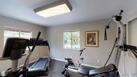 This single-story space showcases a distinctive architectural character defined by prominent exposed wood beam ceilings with warm wood paneling between the beams, lending a mid-century, cabin-inspired aesthetic throughout. The consistent ceiling style unifies the living areas, bedrooms, and kitchen, creating a cohesive and inviting ambiance.

The carpeted living and bedroom areas are generously proportioned, with clean white walls that complement the natural warmth of the wood overhead. One of the main living areas features a full-width sliding glass door that opens directly to a private, fenced outdoor space shaded by a mature tree, allowing natural light to pour in. A bedroom area includes vertical shiplap-style wall paneling and direct access to a walk-in closet.

The kitchen is outfitted with ample white cabinetry, a full suite of white appliances including a refrigerator and electric range, light-toned countertops, and wood-look plank flooring. A window above the sink provides natural light. The bathroom continues the wood-look flooring and features a white vanity with cabinetry, a mirrored medicine cabinet, and a shower-tub combination. Throughout, the space balances functional design with warm, natural materials.