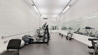 Located at 1710 E Broad St in Richmond, Virginia, this fitness center offers a well-equipped exercise space designed to support a comprehensive workout routine. The room features white-painted exposed brick walls that lend an industrial yet clean aesthetic, complemented by a durable rubber-flooring surface ideal for high-impact activity. Bright fluorescent strip lighting runs along the ceiling, ensuring the space is thoroughly illuminated throughout.

Large wall-mounted mirrors span multiple walls, visually expanding the room and allowing users to monitor their form during exercise. The space is outfitted with a variety of cardio equipment, including treadmills and elliptical machines, as well as a multi-station cable machine for strength training. Free weight options include barbells with weight plates, kettlebells, and a dumbbell rack, providing versatility for a range of fitness goals. A stability ball and workout mat round out the available equipment.

A wall-mounted split air conditioning unit provides climate control, keeping the environment comfortable during intense workouts. The overall design is functional and straightforward, with a clean, minimalist appearance defined by its white walls and efficient layout that maximizes usable floor space for both machine-based and free-weight training.