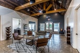 This single-story clubhouse in New Albany, Mississippi, is designed to serve as a welcoming community gathering space. The most striking architectural feature is the dramatic vaulted ceiling clad in warm wood planking and accented by substantial exposed wood beams, which lend a rustic yet refined character to the main lounge area. Ceiling fans suspended from the beams provide comfort throughout the space, while tile flooring runs consistently underfoot.

The main lounge is open and generously proportioned, furnished with leather chairs, a sofa, and a wood coffee table arranged over a patterned area rug. Bold teal-blue accent walls contrast cleanly against crisp white walls and trim. A wall-mounted television and a credenza define a media area off the main lounge, and open shelving units provide decorative display space throughout.

A separate dining or multipurpose room adjacent to the lounge features its own vaulted ceiling, pendant lighting, and windows that invite natural light. A full kitchen is also accessible from the main area, equipped with stainless steel appliances including a refrigerator, range, and microwave, alongside white cabinetry. Arched French doors open directly to an outdoor pool area, reinforcing the clubhouse's role as a social hub for the surrounding community.