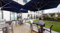 Located at 265 E State St in the heart of downtown Columbus, Ohio, this outdoor courtyard is a well-appointed amenity space designed for relaxation and social gatherings. The space is framed by the surrounding multi-story building façade, creating a semi-enclosed, intimate atmosphere while remaining fully open to the sky above.

The courtyard features a generously sized patio with light-toned pavers providing a clean, durable surface throughout. Multiple dining tables paired with sling-back chairs are arranged across the space, each shaded by large, deep navy market umbrellas that lend a cohesive and sophisticated aesthetic. The bold color of the umbrellas contrasts elegantly with the neutral tones of the building and hardscape.

A section of lush artificial turf adds a verdant, park-like quality to the space, softening the overall look and offering a visual break from the hardscape. Raised planter beds and potted tropical greenery introduce additional natural elements, enhancing the outdoor ambiance. A designated lounge area with seating is also present, offering a more relaxed alternative to the dining arrangements.

The space is enclosed by sleek metal railings and benefits from direct access via glass-paneled doors leading to the interior. The surrounding urban skyline is visible beyond the building perimeter, grounding this tranquil retreat firmly within the vibrant Columbus cityscape.