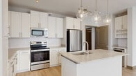 This well-appointed single-floor residence offers a thoughtfully designed layout that balances comfort and functionality. The open-concept kitchen serves as a centerpiece, featuring white shaker cabinetry, a classic subway tile backsplash, stainless steel appliances including a refrigerator, electric range, and over-the-range microwave, and a spacious island with a stone countertop and undermount sink. Globe pendant lights overhead lend a modern touch, while light hardwood-style flooring extends seamlessly into the adjoining living area, which benefits from abundant natural light through multiple windows and a ceiling fan. A dedicated wet bar nook with built-in shelving adds extra functionality near the main living space. The bedrooms feature plush carpeting, ceiling fans, and generous natural light from well-placed windows. The bathroom is cleanly finished with white tile surrounds on a full soaking tub and shower combination, complemented by a vanity with stone countertops. A dedicated laundry area houses an in-unit washer, while a walk-in closet with wire shelving provides ample storage. Throughout, clean white walls, recessed lighting, and a crisp, neutral palette create a bright and contemporary ambiance. A long, well-lit corridor with light wood flooring connects the spaces with ease.