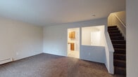 This two-floor residence offers a practical and functional layout across its living spaces. The main living area is generously sized, finished with wall-to-wall carpeting in a neutral tone, and painted in soft gray-white hues that lend a clean, understated ambiance. A baseboard heating system runs along the perimeter, and a large window fitted with vertical blinds allows natural light to fill the space. An open pass-through connects the living area to the galley-style kitchen, which features warm oak cabinetry on both sides, light countertops, a double-basin stainless steel sink, and white appliances including a gas range and refrigerator. The kitchen flooring is a light-toned vinyl, offering easy maintenance. Bedrooms are found on the upper level and showcase warm hardwood flooring in a honey oak finish, complemented by baseboard heaters and windows with vertical blinds that bring in natural light. The bathroom includes an extended vanity countertop with an integrated sink, a large wall mirror, and a tiled tub-and-shower combination. Throughout, the space maintains a neutral color palette and straightforward design, providing a versatile and move-in-ready environment.