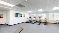 Located at 21620 Dracaea Avenue in Moreno Valley, CA, this fitness center offers a well-organized and fully equipped exercise space designed to support a comprehensive workout routine. The single-floor facility features a clean, modern aesthetic with crisp white walls, dark baseboards, and durable speckled carpet flooring suited for active use. A drop-panel ceiling with recessed LED lighting ensures the space is consistently and evenly lit, while multiple windows fitted with horizontal blinds allow natural light to supplement the interior illumination and provide views of the surrounding outdoors. The open layout accommodates a wide variety of equipment, including two Spirit-brand treadmills, an elliptical trainer, a stationary spin bike, and a recumbent-style cycle. Strength training needs are addressed through a multi-function cable machine, a dedicated leg press/seated machine, an adjustable weight bench, a flat utility bench, and a full rack of dumbbells. A stability ball rounds out the functional fitness offerings. A wall-mounted flat-screen television is positioned for easy viewing during cardio sessions, accompanied by motivational artwork. The entrance features a glass-panel door with an "Altitude" branded mat, adding a polished, community-fitness feel to the space.