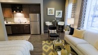 This thoughtfully designed studio offers an open-concept floor plan that combines the living, dining, and kitchen areas into one cohesive, functional space. The kitchen is outfitted with dark espresso cabinetry, stainless steel appliances including a full-size refrigerator, and light stone countertops, providing a striking contrast against the rich cabinetry. Durable hardwood-style flooring runs throughout, adding warmth and continuity across the entire space. The living area is anchored by a neutral sofa and complemented by bold yellow and geometric-patterned accents, including area rugs, throw pillows, and floor-length curtains that frame a bright window, allowing natural light to fill the room. A compact bistro-style dining set with white bar stools serves as the dining area, perfectly scaled for the space. The accent wall in deep charcoal gray adds depth and a modern aesthetic, while framed artwork contributes to the curated, contemporary feel. Overall, the space balances style and practicality, making efficient use of its open layout to create a comfortable and visually cohesive living environment.
