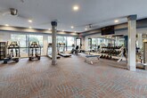 This expansive fitness center is thoughtfully designed to accommodate a full range of workout needs within a single, open floor plan. The space features a neutral, patterned carpet throughout and is anchored by structural columns that define distinct workout zones without disrupting the overall flow of the room. Large windows line the perimeter walls, flooding the interior with natural light and providing views of the surrounding greenery, creating an inviting and energizing atmosphere.

The cardio zone is well-stocked with treadmills, ellipticals, and stationary bikes from TRUE Fitness, all positioned along the windowed walls to maximize natural light during workouts. Multiple wall-mounted flat-screen TVs are positioned throughout the space for entertainment. A dedicated strength training area features a comprehensive rack of dumbbells displayed on mirrored walls, along with weight benches and a variety of selectorized cable and resistance machines for targeted muscle training.

An adjoining studio room with mirrored walls and a ballet barre offers space for stretching, yoga, or group fitness activities. A bold blue accent wall near the entrance holds stability balls in multiple sizes and a water fountain station with a bottle-filling feature, adding both color and functionality. Recessed lighting supplements the natural light throughout, ensuring the space remains well-lit at all times.