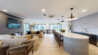 Located in Woodinville, WA, this thoughtfully designed single-floor community space offers an open, flowing layout that seamlessly connects a kitchen, living room, and dining area. The kitchen is a standout centerpiece, anchored by a large oval island clad in mosaic tile and topped with a smooth stone countertop, complemented by a striking geometric tile backsplash, dark cabinetry, stainless steel appliances including a wine refrigerator, and a trio of sculptural pendant lights overhead. Light wood-tone flooring runs throughout, lending warmth and continuity across the space. The living area features a linear gas fireplace set within a textured tile surround, a wall-mounted display, and large floor-to-ceiling windows that frame lush Pacific Northwest views and invite abundant natural light. A glass door opens directly onto a spacious outdoor terrace finished with large-format tile, offering seating areas ideal for gathering while overlooking the surrounding rooftops and rolling greenery. A dedicated game room adds a distinctive touch, with sage-green walls adorned with a collection of decorative wine barrel medallions and a custom round gaming table at its center. The overall ambiance is contemporary and refined, balancing a welcoming social atmosphere with high-end finishes throughout.