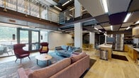 This contemporary two-story space is defined by its industrial-modern aesthetic, combining exposed ductwork, dark structural beams, and sleek linear lighting with warm wood tones and carefully curated furnishings. The main level centers around an expansive open-concept lounge area featuring double-height ceilings that amplify the sense of volume. Comfortable seating arrangements — including a blue sofa, deep burgundy accent chairs, and a round wood coffee table — are anchored by patterned area rugs over light hardwood flooring. A dining area with bold yellow chairs adds a vibrant contrast nearby, while glass doors lead to an exterior balcony, inviting natural light and outdoor views.

The kitchen areas are thoroughly equipped for serious use, featuring light wood cabinetry, geometric tile backsplashes, quartz countertops, stainless steel appliances including a professional-grade refrigerator, built-in cooktop, microwave, and a large central island. A secondary kitchenette provides additional prep space with similar finishes.

A central staircase with polished stainless steel and glass railings connects the floors, crowned by a dramatic multi-pendant chandelier that cascades dramatically through the open stairwell. The upper mezzanine level overlooks the main lounge below, reinforcing the space's open, connected character throughout.