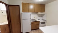 This single-floor unit offers a straightforward, functional layout across several rooms. The kitchen is equipped with warm oak-toned cabinetry extending to the ceiling, providing ample storage. White appliances include a top-freezer refrigerator, an electric coil range with oven, and a single-basin sink set beneath a range hood. Tile flooring runs throughout the kitchen and bathroom, offering durability in high-use areas.

The living area and bedroom spaces feature wall-to-wall carpet in neutral tones, contributing a quieter, softer atmosphere. Walls are painted white throughout, keeping the interior bright and neutral. Baseboard heating units run along the lower walls, serving as the primary heat source. Each carpeted room includes at least one window, allowing natural light to filter in.

The bathroom is compact and utilitarian, featuring a full-size soaking tub with a tub/shower combination fitted with a curtain rod. Tile flooring continues here, coordinating with the kitchen. Wood-tone interior doors with brass hardware appear consistently throughout the unit, providing a cohesive, classic finish. Overall, the space presents a clean, no-frills interior suited for practical everyday living.