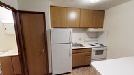 This single-floor unit offers a straightforward, functional layout across several rooms. The kitchen is equipped with warm oak-toned cabinetry extending to the ceiling, providing ample storage. White appliances include a top-freezer refrigerator, an electric coil range with oven, and a single-basin sink set beneath a range hood. Tile flooring runs throughout the kitchen and bathroom, offering durability in high-use areas.

The living area and bedroom spaces feature wall-to-wall carpet in neutral tones, contributing a quieter, softer atmosphere. Walls are painted white throughout, keeping the interior bright and neutral. Baseboard heating units run along the lower walls, serving as the primary heat source. Each carpeted room includes at least one window, allowing natural light to filter in.

The bathroom is compact and utilitarian, featuring a full-size soaking tub with a tub/shower combination fitted with a curtain rod. Tile flooring continues here, coordinating with the kitchen. Wood-tone interior doors with brass hardware appear consistently throughout the unit, providing a cohesive, classic finish. Overall, the space presents a clean, no-frills interior suited for practical everyday living.