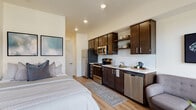 Located at 6347 SW Canyon Ct in Portland, Oregon, this single-floor studio unit offers a well-designed open-concept layout that efficiently combines living, sleeping, dining, and cooking areas into one cohesive space. Light wood-style flooring runs throughout, lending warmth and visual continuity across every zone.

The kitchen is positioned along one wall and is outfitted with dark espresso cabinetry, open shelving, a white subway tile backsplash, light countertops, and a full suite of stainless steel appliances, including a refrigerator, gas range, microwave, and dishwasher. The living area sits adjacent to the kitchen and benefits from generous natural light provided by large floor-to-ceiling windows and a glass-paned door, offering views of the surrounding neighborhood. The sleeping area is tucked toward the rear of the unit, creating a sense of separation within the open floor plan.

A private hallway leads to the full bathroom, which features the same wood-style flooring, a dark vanity with a white countertop, a glass-enclosed shower, and clean white walls. In-unit laundry with a washer and dryer is also accessible off this corridor, adding everyday convenience to the space.
