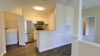 This single-floor space showcases a well-appointed kitchen that serves as a central feature of the layout. The kitchen is designed with clean white cabinetry that extends to the upper walls, providing ample storage. Granite countertops with subtle veining add a refined touch, complementing the full suite of stainless steel appliances, which includes a refrigerator, over-the-range microwave, and an electric range with matching finishes.

A peninsula with a white base extends from the main kitchen area, creating a natural divide between the kitchen and the adjacent living space. This layout offers both a functional workspace and a casual gathering point. The dark hardwood flooring runs throughout, providing a strong visual contrast against the white walls and cabinetry, lending the space a modern and polished aesthetic.

The walls are painted in a warm neutral tone, enhancing the welcoming ambiance. Flush-mounted ceiling lighting provides consistent illumination throughout the area. The open sight lines between the kitchen and the connected space contribute to an airy, functional flow that makes the layout feel cohesive and practical.