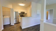 This single-floor space showcases a well-appointed kitchen that serves as a central feature of the layout. The kitchen is designed with clean white cabinetry that extends to the upper walls, providing ample storage. Granite countertops with subtle veining add a refined touch, complementing the full suite of stainless steel appliances, which includes a refrigerator, over-the-range microwave, and an electric range with matching finishes.

A peninsula with a white base extends from the main kitchen area, creating a natural divide between the kitchen and the adjacent living space. This layout offers both a functional workspace and a casual gathering point. The dark hardwood flooring runs throughout, providing a strong visual contrast against the white walls and cabinetry, lending the space a modern and polished aesthetic.

The walls are painted in a warm neutral tone, enhancing the welcoming ambiance. Flush-mounted ceiling lighting provides consistent illumination throughout the area. The open sight lines between the kitchen and the connected space contribute to an airy, functional flow that makes the layout feel cohesive and practical.