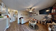 This well-organized single-story home offers a thoughtfully designed layout comprising a combined kitchen and living room, one bedroom, a dedicated office, and a full bathroom. The living room serves as the heart of the home, featuring rich wood-look flooring, a ceiling fan with light fixture, and a striking floor-to-ceiling stone fireplace that anchors the space with rustic character. A sliding glass door beside the fireplace allows natural light to filter in and provides outdoor access. The combined living and kitchen area flows seamlessly, with the galley-style kitchen offering light wood cabinetry, stainless steel appliances including a full-size refrigerator and range, and a window that brings in additional natural light. The bedroom is a comfortable retreat with wood-look flooring, a ceiling fan, and direct access to the bathroom. The bathroom features a warm wood vanity with a granite-style countertop and a well-lit mirror. The dedicated office space provides a functional work area with a window fitted with blinds for light control. Throughout the home, textured ceilings add architectural detail, and the consistent wood-look flooring ties each room together for a cohesive, inviting aesthetic.