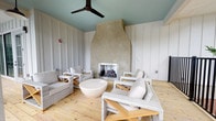 Located at 1235 Ashley Gardens Blvd in Charleston, South Carolina, this single-floor community space offers a warm and welcoming environment with a distinctly coastal character. The centerpiece of the lounge is a striking plaster-surround fireplace, rendered in a smooth, earthy tone that draws the eye and anchors the room with a sense of architectural elegance. Light pine hardwood flooring runs throughout, lending warmth and a natural texture underfoot. The walls are finished in crisp white board-and-batten paneling, creating a clean, classic backdrop that complements the relaxed aesthetic. A teal-painted ceiling adds a subtle pop of coastal color overhead, while a sleek dark ceiling fan ensures comfortable airflow year-round. The seating arrangement features woven rattan and teak lounge chairs paired with plush neutral cushions, all centered around a smooth, rounded white coffee table — an inviting setup ideal for gathering and conversation. The overall ambiance is light, airy, and thoughtfully designed, blending Southern charm with contemporary coastal style to create a comfortable and visually cohesive communal retreat.