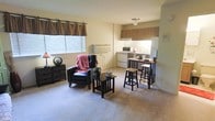 This single-floor unit offers an open-concept layout that seamlessly connects the living area and kitchen, creating a practical and comfortable living environment. The living space is finished with soft carpeting and features warm, neutral-toned walls that lend a calm, welcoming ambiance. A large window dressed with decorative valance curtains allows natural light to fill the room, brightening the space throughout the day. A wall-mounted climate control unit ensures year-round comfort.

The kitchen area is efficiently designed with wooden cabinetry in a warm medium-brown tone, providing ample storage. White appliances, including a refrigerator, range, and overhead range hood, are neatly arranged along the kitchen wall. A compact breakfast bar with bar stools serves as a casual dining and gathering spot, making excellent use of the available space.

The living area accommodates a recliner chair and side tables, offering a relaxed seating arrangement. The open flow between the kitchen and living spaces makes the unit feel airy and connected, ideal for convenient day-to-day living. Overall, the unit combines functional design with a cozy atmosphere, making it a practical and comfortable single-floor residence.