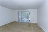 This single-floor residence presents a straightforward and functional layout designed for comfortable everyday living. The living areas are finished with wall-to-wall neutral-toned carpeting, lending a warm and cohesive feel throughout the space. Bright white walls create a clean, open atmosphere, and a large sliding glass door fitted with vertical blinds allows natural light to fill the main living area while providing access to the outdoors and views of surrounding greenery.

The floor plan flows openly between the living and adjoining rooms, with multiple doorways connecting the various spaces. A ceiling fan is present in one of the main living areas, adding both comfort and a functional accent. The kitchen is equipped with warm-toned wood cabinetry above and below the counters, a double stainless steel sink, white countertops, and a full-size black refrigerator, providing ample storage and workspace for everyday meal preparation. A stove is also present in the kitchen area.

The bathroom features a classic white tile surround enclosing a full bathtub with an overhead showerhead and chrome fixtures, complemented by clean white walls that enhance the sense of brightness. The overall design of the space is practical and well-maintained, offering a neutral canvas well-suited for personalization.