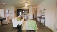 This single-floor residence showcases an open-concept layout that connects the living and kitchen areas into one cohesive, functional space. The living area is comfortably sized, featuring rich, dark hardwood-style flooring that runs throughout, anchoring the space with warmth and continuity. A ceiling fan with integrated lighting occupies the center of the room, providing both comfort and illumination. The seating area includes a cream-toned sofa adorned with green and brown accent pillows, complemented by a light upholstered ottoman and a glass-top coffee table, creating a relaxed yet polished atmosphere.

The kitchen, directly accessible from the living space, is equipped with dark espresso cabinetry contrasted by light countertops, stainless steel appliances including a refrigerator and microwave, and a peninsula with bar-height seating that serves as a natural divider. Pendant lighting above the peninsula adds a refined touch. A narrow console table along one wall, topped with framed artwork and a decorative bowl, contributes an elegant accent. Hallways on either side suggest additional private rooms beyond the main living area, completing this well-appointed and thoughtfully designed home.