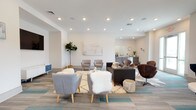 Located at 300 Bellamy Ave in Conway, South Carolina, this single-floor community amenity space is thoughtfully designed to foster social interaction and relaxation. The open-concept layout seamlessly connects multiple distinct zones, all unified by a bright, contemporary aesthetic with crisp white walls, recessed lighting, and a combination of wood-look and patterned carpet flooring in neutral tones accented with teal.

The expansive lounge area serves as the heart of the space, furnished with an eclectic mix of seating including a dark sofa, swivel chairs in rich brown leather, taupe accent chairs, and plush ottomans arranged around a central area rug. Multiple flat-screen televisions are mounted throughout, enhancing the entertainment experience. A vintage arcade game adds a playful touch to one corner of the lounge.

A dedicated bar and kitchenette area features gray shaker-style cabinetry, granite countertops, stainless steel appliances including a beverage refrigerator and dishwasher, and a pass-through window. An adjacent high-top communal table with black metal stools provides a casual dining and gathering spot, complemented by a wall-mounted TV and textured blue tile backsplash.

Large glass double doors flood the space with natural light and provide direct access to an outdoor balcony, while coastal-inspired artwork and lush indoor plants contribute to the welcoming, resort-like ambiance throughout.