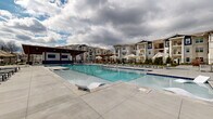 This two-floor residential community is centered around a generously scaled outdoor amenity area designed for leisure and social interaction. The centerpiece is a large, resort-style swimming pool with crystal-clear blue water, multiple entry points via stainless steel handrails, and built-in in-water lounge chairs that allow residents to relax directly in the pool. The surrounding concrete pool deck provides ample room for sunbathing and is lined with umbrella-shaded seating areas for additional comfort.

Adjacent to the pool, a covered outdoor pavilion serves as a social hub. Constructed with heavy timber columns and a warm wood-paneled ceiling with recessed lighting, the pavilion features a built-in bar-height counter with wall-mounted brackets, ideal for outdoor dining and entertaining. A mounted flat-screen TV adds a recreational element to the space.

Beyond the pavilion, an expansive open-air patio extends the usable outdoor area, furnished with high-top tables and chairs. A pergola-shaded seating zone provides a quieter retreat surrounded by manicured landscaping. The clubhouse building features large floor-to-ceiling glass doors, prominent columns, and a clean white board-and-batten exterior, creating an inviting and modern architectural focal point for the community.